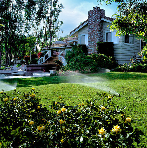 Sprinkler watering a lawn with a house and trees in the background