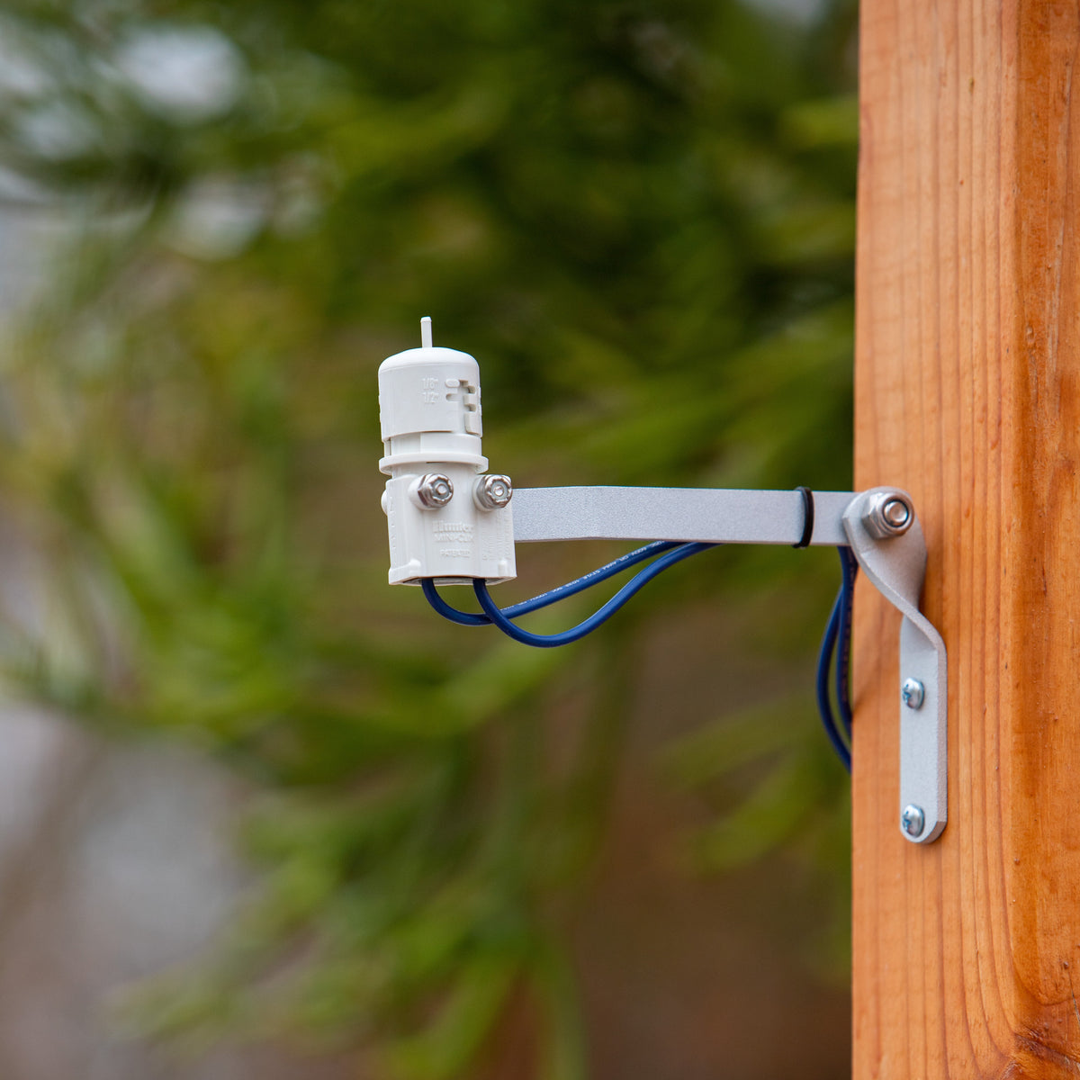 A white outdoor mini-clik rain sensor with a silver mounting bracket and blue wiring, mounted on a wooden post with a blurred green background.