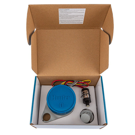 Open cardboard box containing a blue device, wires, and a bottle on a white background