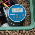 Hunter Node-100 irrigation timer with digital display and buttons, placed on a bed of small stones.