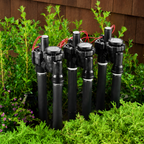 Set of black garden sprinklers in a garden setting with green plants and a wooden fence.