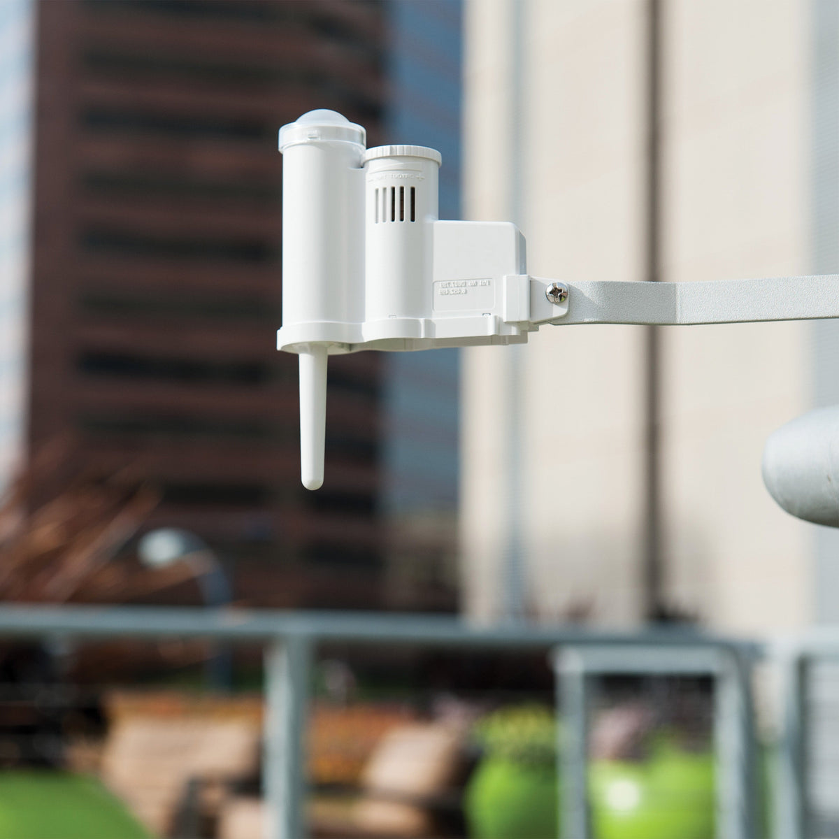 A white outdoor solar sync sensor mounted on a metal bracket, shown against a blurred city background with buildings and sky.
