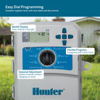 Hunter X2 easy dial programmable irrigation controller shown with feature callouts on a blurred outdoor lawn background. The controller has a gray housing, blue front panel, digital display, directional buttons, and a large rotary dial.