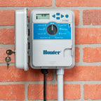 Hunter X-Core irrigation controller with dial interface and LCD display mounted on brick wall, grey and blue casing.