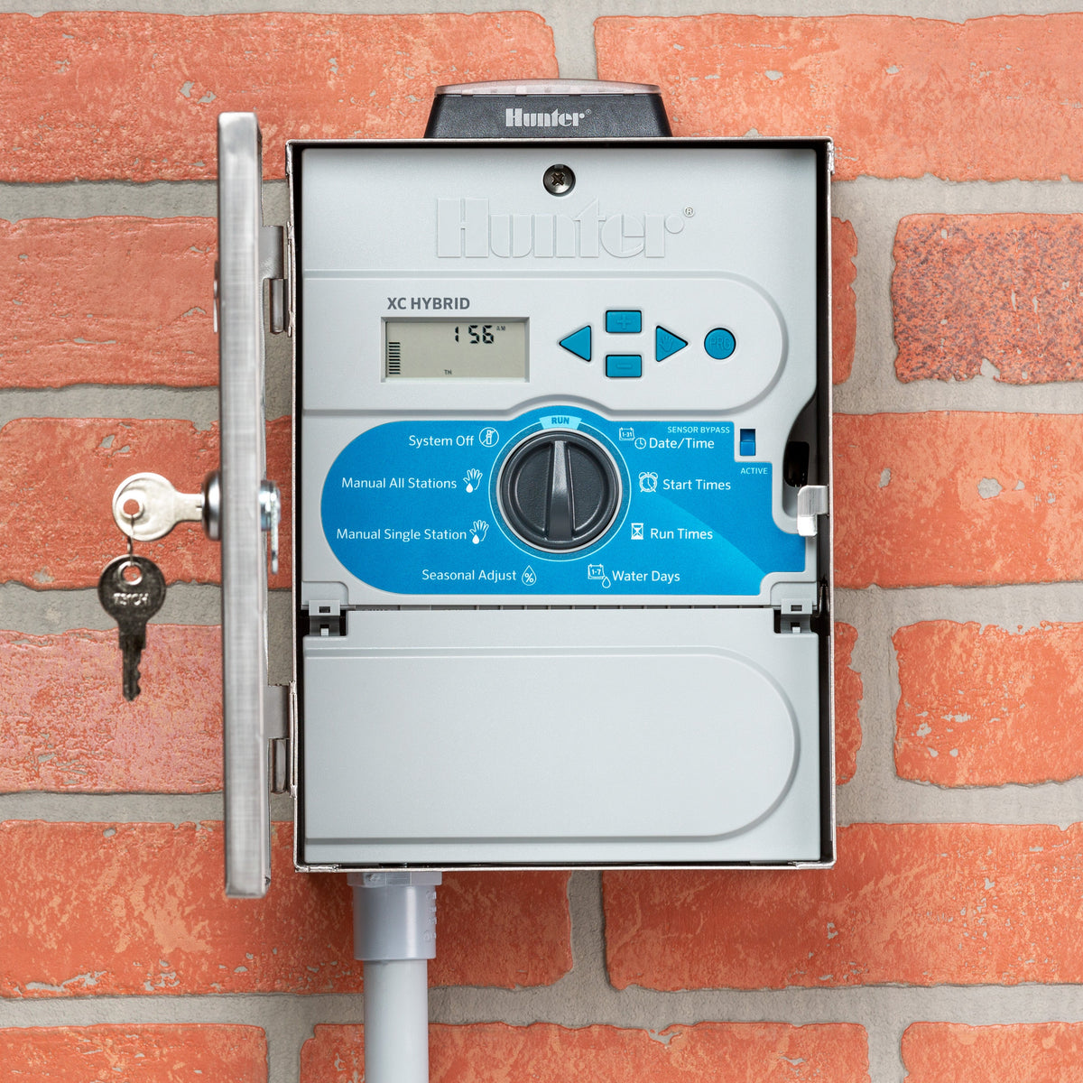 Gray Hunter X-Core solar-powered irrigation controller with digital screen and blue control dial mounted on brick wall.